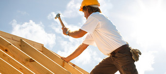 Dachdecker mit gelbem Schutzhelm beim Befestigen von Konstruktionsvollholz (KVH) auf einem Dachstuhl unter strahlendem Himmel.
