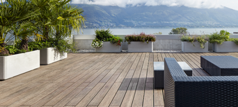 Große Terrasse mit Bambus-Holzdielen, modernen Rattanmöbeln, Pflanzen in Betonkübeln und Blick auf einen See mit Bergen.