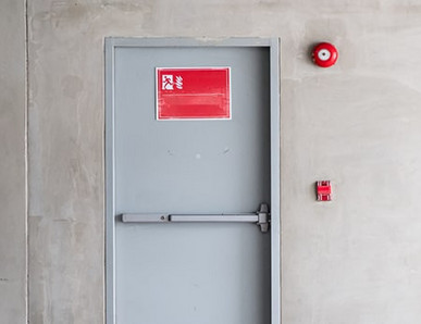 Graue Brandschutztür mit Fluchtwegsymbol und roter Alarmglocke.